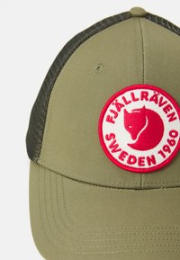 Olīvzaļa cepure ar melnu sietu aizmugurē un apaļu sarkanu un baltu Fjällräven Zviedrija 1960 logo uzlīmi ar lapsas galvu.
