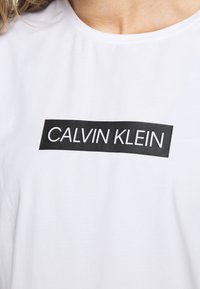 Biała bawełniana koszulka z czarnym prostokątnym logo z napisem "CALVIN KLEIN" w grubych białych literach na piersi.