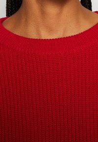 Gros plan sur le cou et le haut de la poitrine d'une personne portant un pull rouge en maille avec un col rond.