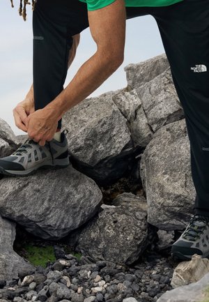 Person, die schwarze Hose und grünes Hemd trägt, bindet grauen Wanderschuh, während sie auf großen grauen Felsen im Freien steht.