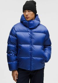 Sinine puff-jakk kõrge kraega, läikiv materjal, horisontaalne tikandimuster, luku sulgemine ja must müts. Reguleeritav kapuuts nuppudega.