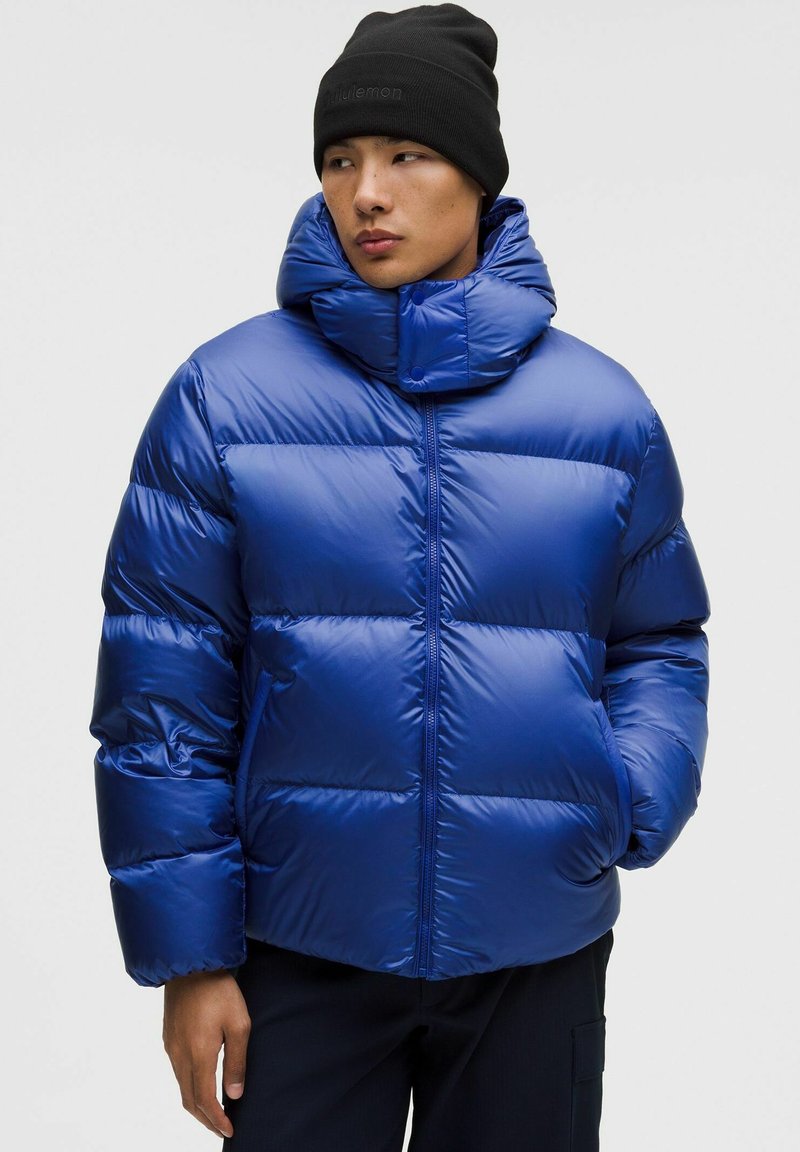 Sinine puff-jakk kõrge kraega, läikiv materjal, horisontaalne tikandimuster, luku sulgemine ja must müts. Reguleeritav kapuuts nuppudega.