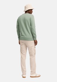 Sudadera verde clara de manga larga con puños de canalé, combinada con pantalones cargo crema. Lleva un sombrero de cubo beige y zapatillas blancas.