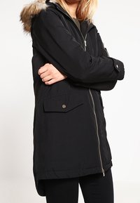 Černá parka bunda s kapucí lemovanou umělou kožešinou, předním zipem, dvěma velkými bočními kapsami a mírně delším zadním lemem. Hladká textura.