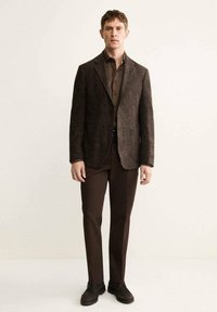 Blazer in tweed marrone con rever a punta, abbinato a una camicia e pantaloni marrone scuro. Accompagnato da scarpe marrone scuro. Sfondo semplice.