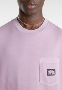 T-shirt in cotone viola chiaro con colletto a girocollo, maniche corte e taschino sul petto con logo Vans nero. Texture liscia, design casual.