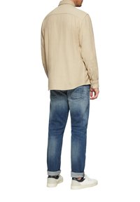 Beige Langarmhemd mit strukturierter Oberfläche und Rückenpasse, kombiniert mit hellblauen Jeans mit leichter Ausbleichung und hochgekrempelten Ärmeln. Weiße Sneaker.
