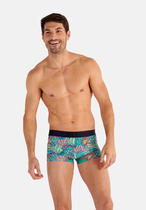 Homme souriant torse nu portant des boxers à motifs tropicaux colorés avec une ceinture sombre sur un fond clair uni.