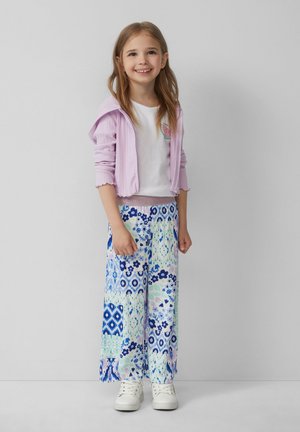 Hellblauer, gerippter Hoodie, weißes T-Shirt mit Wassermelonen-Logo und weite, gemusterte Hosen in blau-grünen Blumenmotiven. Weiße Sneaker.