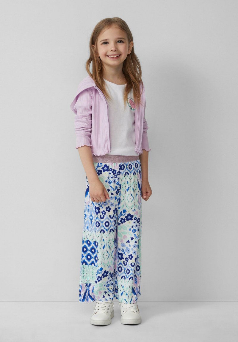 Hellblauer, gerippter Hoodie, weißes T-Shirt mit Wassermelonen-Logo und weite, gemusterte Hosen in blau-grünen Blumenmotiven. Weiße Sneaker.