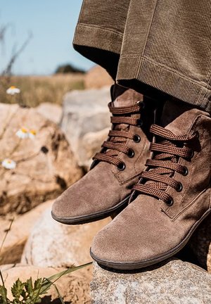 Bottines en daim marron avec lacets, portées avec un pantalon en velours côtelé marron, reposant sur des rochers dans un cadre naturel en plein air avec des fleurs sauvages.