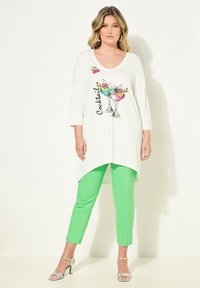 Tunica bianca con un design grafico di cocktail, abbinata a pantaloni verdi vivaci. La tunica presenta un scollo a V e un orlo asimmetrico. Sandali argentati.