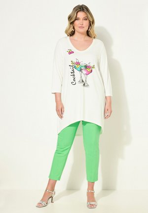 Witte tuniek met een grafisch cocktailontwerp, gecombineerd met levendig groene broeken. De tuniek heeft een V-hals en een hoge-laag zoom. Zilveren sandalen.