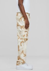 Beige camo cargo pants made of cotton, featuring side pockets and a loose fit. Paired with white sneakers, viewed from the side.