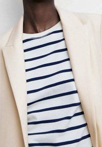 Světle béžový sako přes bílo-námořnický pruhovaný košili. Sako má strukturovaný design, zatímco košile je s vodorovnými pruhy.