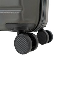 Primer plano de la esquina de una maleta negra con superficie texturizada y dos ruedas dobles giratorias sobre fondo blanco.