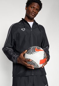 Sort træningsjakke med hvide accentlinjer, lynlås foran og snoretræk i hætten; holder en mønstret fodbold med orange og sorte detaljer.