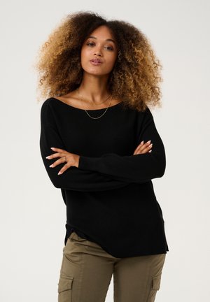 Pulover negru tricotat cu un decolteu off-the-shoulder, completat cu mâneci mansetate, purtat peste pantaloni cargo verzi măslinii.