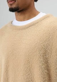 Beige, zacht gebreide trui met een brede halslijn en losse pasvorm, gedragen over een wit shirt met ronde hals, met een zachte, gestructureerde stof.