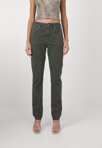 Pantalones verde oliva con un diseño ajustado, estilo de cinco bolsillos, pierna recta y una textura sutil. Combinados con tacones blancos con tiras.