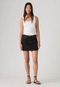 Νεαρή γυναίκα που στέκεται, φοράει λευκό αμάνικο τοπ, μαύρη μίνι φούστα από τζην και μαύρα σανδάλια με λουράκια, μπροστά σε απλό γκρι φόντο.