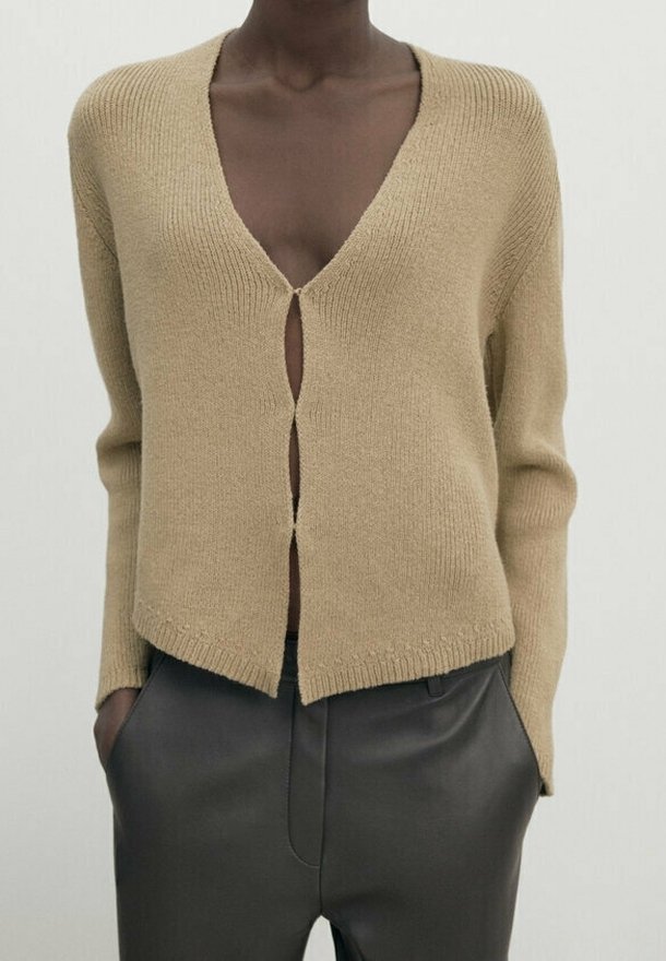 Beige knit cardigan with a deep V-neck and a front opening, featuring ribbed texture and long sleeves. Paired with dark trousers.