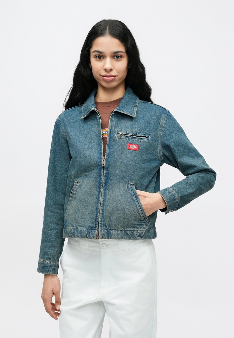 Giovane donna con lunghi capelli scuri che indossa una giacca di jeans blu e pantaloni bianchi, in piedi contro uno sfondo chiaro e uniforme.