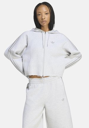 Donna con capelli intrecciati che indossa una felpa con cappuccio grigio chiaro con zip e pantaloni della tuta abbinati, in piedi con le mani sui fianchi contro uno sfondo bianco.