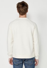 Felpa color crema con un motivo testurizzato a piqué, polsini e orlo a coste. Presenta un collo rotondo e maniche raglan.