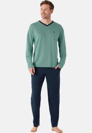 Mann steht barfuß, trägt ein grünes Langarmshirt mit V-Ausschnitt und dunkelblaue Hose, blickt leicht seitlich und hat die Hände in den Taschen.