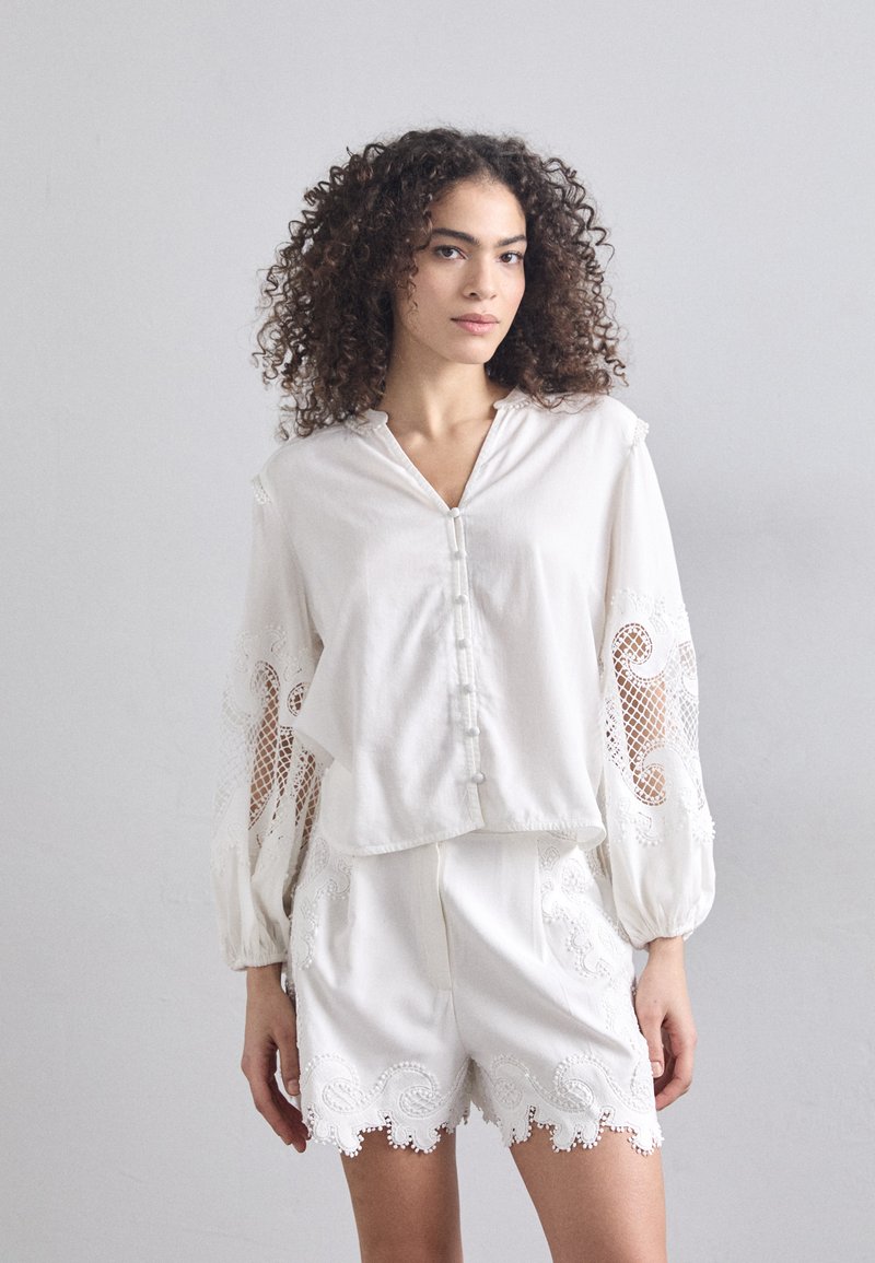 Vit blus med långa, puffiga ärmar och spetsdetaljer, tillsammans med vita shorts som har scallopade kanter och broderade accenter.
