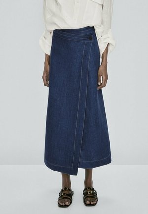 Jupe portefeuille en denim bleu foncé avec fermeture par bouton, portée avec un chemisier blanc à manches longues et des sandales noires à boucle sur une personne debout.