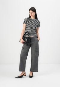 T-shirt risca preta e branca, calças cargo cinzas com bolsos e saltos slingback pretos. A segurar uma pequena mala preta com uma alça trançada.