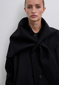 Abrigo de lana negro con un gran cuello, que presenta un cierre de botones y tela drapeada. Suave textura y un silueta sobredimensionada realzan su diseño.