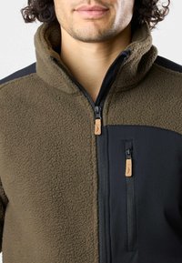 Fleecejacke mit einem braunen strukturierten Körper und schwarzen glatten Ärmeln. Bedruckter Front-Reißverschluss mit kontrastierenden Hardware-Akzenten. Hoher Kragen.