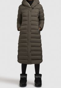 Gestepptes olivgrünes langes Parka mit Kapuze, mit Knopfverschluss, Seitentaschen und einer strukturierten Stoffoberfläche. Tragen über schwarzen Leggings und Stiefeln.