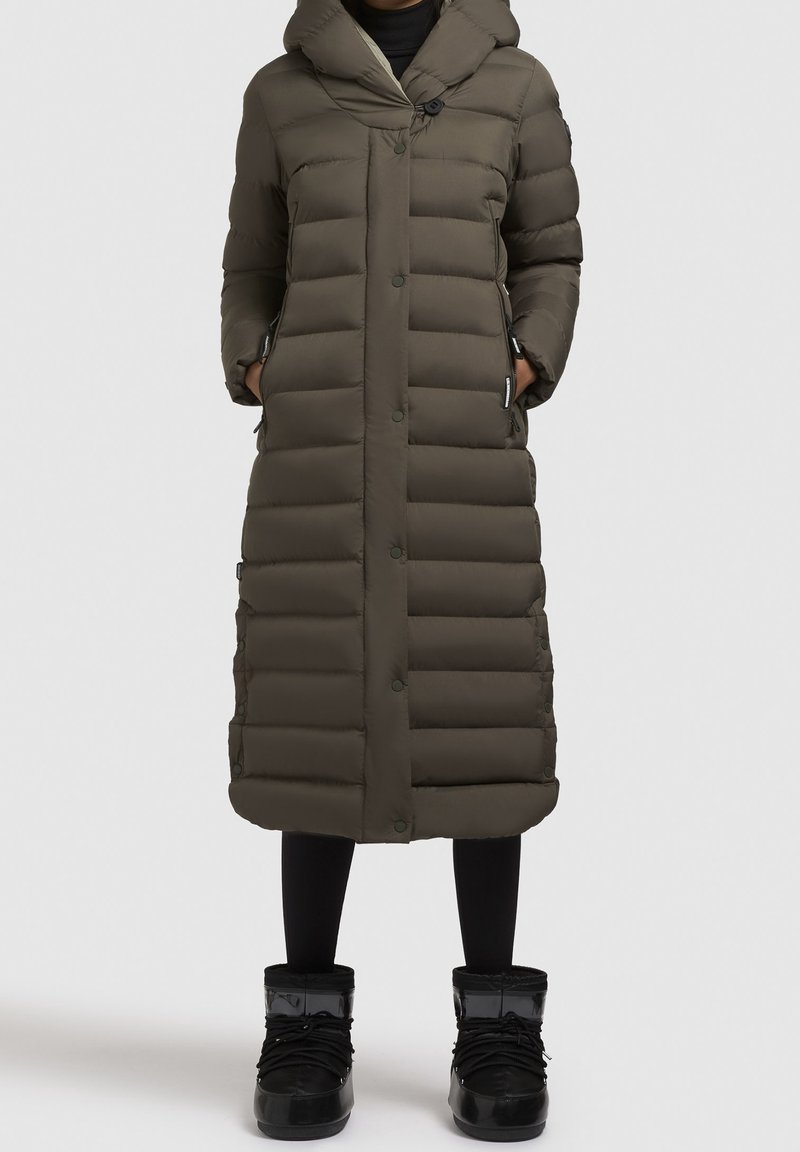 Gestepptes olivgrünes langes Parka mit Kapuze, mit Knopfverschluss, Seitentaschen und einer strukturierten Stoffoberfläche. Tragen über schwarzen Leggings und Stiefeln.