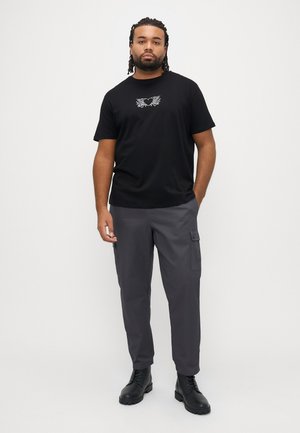 Fekete, rövid ujjú póló fehér szív- és szárnygrafikával, szürke cargo nadrággal és fekete, fűzős csizmával párosítva.