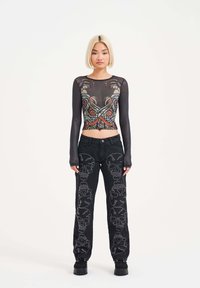 Top negro transparente de manga larga con diseño gráfico colorido; jeans negros de tiro alto con bordado de calaveras blancas a lo largo de las piernas. Calzado negro.