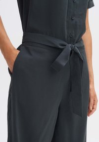 Dunkelgrauer Jumpsuit aus glattem Stoff, mit einem gebundenen Taillengürtel und kurzen Ärmeln, einem runden Ausschnitt und Knopfdetails.