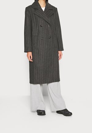 Femme portant un long manteau croisé gris foncé à fines rayures, un pantalon large gris clair et des baskets noires sur un fond uni.