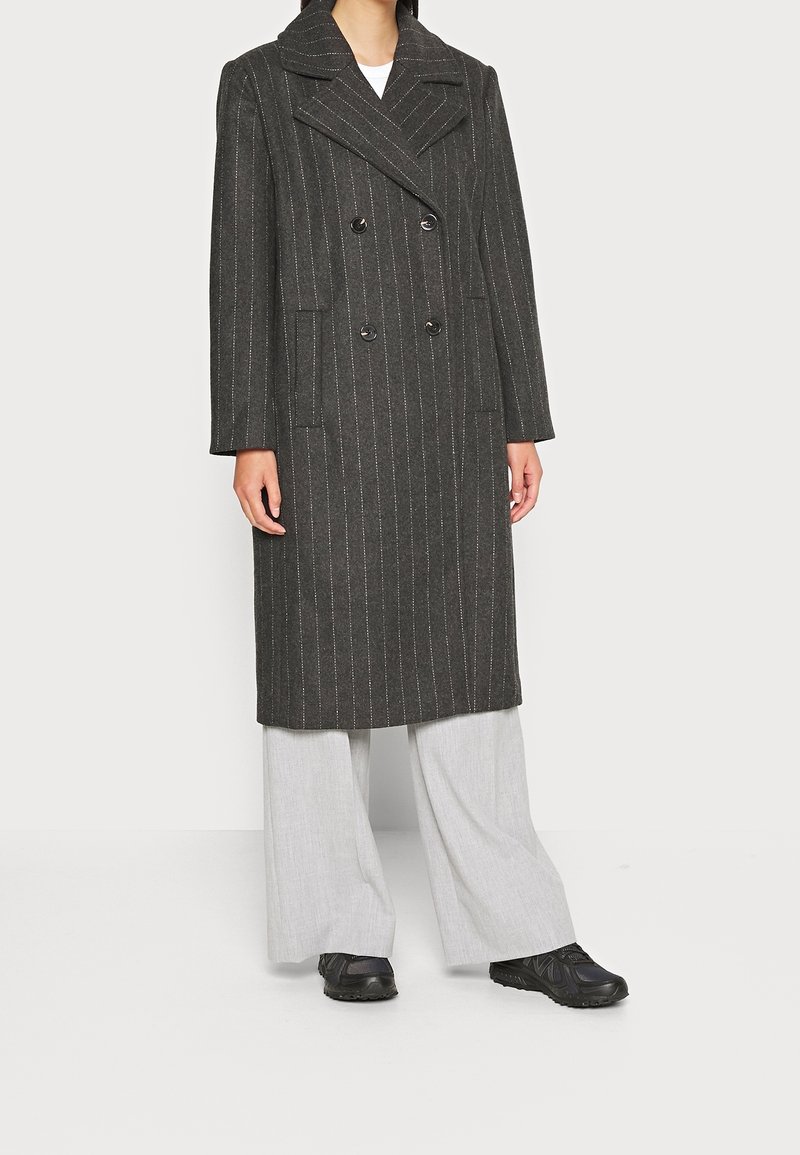 Femme portant un long manteau croisé gris foncé à fines rayures, un pantalon large gris clair et des baskets noires sur un fond uni.