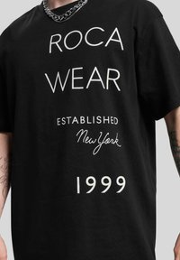 Melnā kokvilnas T-krekliņa ar izteiksmīgu baltu tekstu: "ROCA WEAR", "ESTABLISHED", "New York" un "1999" dažādās fontu un izmēru kombinācijās. Redzama ķēdes kaklarota.