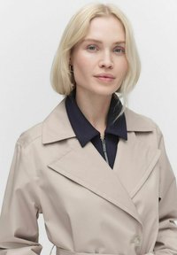 Beżowy trencz z dużym kołnierzem i granatową, zapinaną na zamek wewnętrzną warstwą. Tkanina wydaje się gładka z subtelnym połyskiem. Dopasowany krój z guzikami.