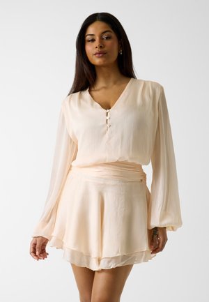 Kobieta o długich ciemnych włosach, ubrana w jasnobeżową, długorękawową bluzkę z guzikami oraz pasującą warstwową spódnicę, stojąca na jednolitym tle.