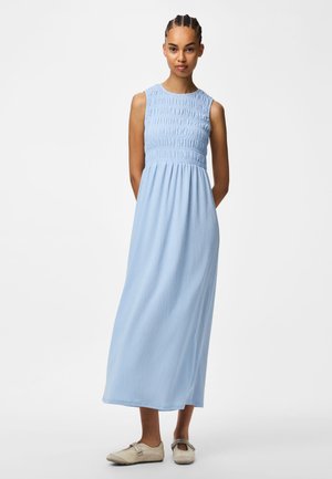 Γυναίκα που στέκεται με τα χέρια πίσω από την πλάτη, φοράει αμάνικο ανοιχτό μπλε midi φόρεμα και κρεμ παπούτσια slip-on σε λευκό απλό φόντο.