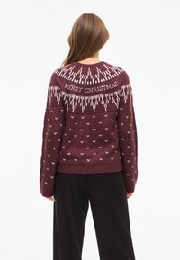Burgundy knit sweater with white "MERRY CHRISTMAS" text, geometric patterns, and heart motifs. Long sleeves and a ribbed hem.