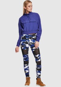 Kuttet lilla jakke med en frontlomme, kombinert med blå, svart og hvit camo tights med gjennomsiktige detaljer og brune snørestøvler.
