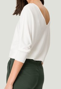 Haut blanc à manches longues avec un dos en V et des poignets côtelés, associé à un pantalon ajusté vert foncé, mettant en avant la texture et le contraste.