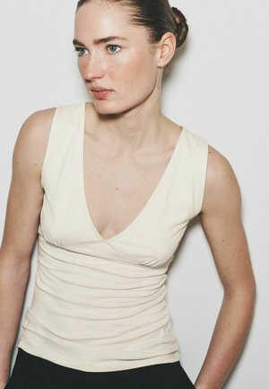 Junge Frau mit blauen Augen und geflochtenem Haar trägt ein ärmelloses cremefarbenes Top mit tiefem V-Ausschnitt, stehend vor einem einfarbigen hellen Hintergrund.